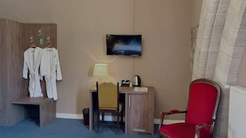 A hotel room with a wooden wardrobe, a desk with a lamp, a television, a red chair, and a small trash bin.