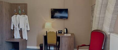 A hotel room with a wooden wardrobe, a desk with a lamp, a television, a red chair, and a small trash bin.