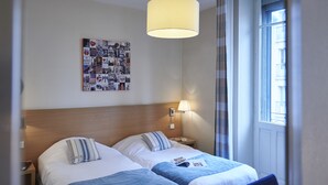 Habitación de hotel con dos camas, una composición de arte en la pared, una lámpara y una ventana con cortinas.