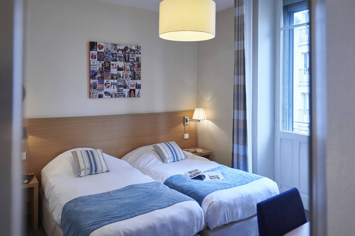 A hotel room with two beds, a wall art collage, a lamp, and a window with curtains.