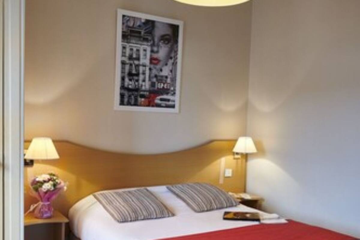 A hotel room with a bed, two bedside lamps, a red throw blanket, a framed picture on the wall, and a blue carpet.
