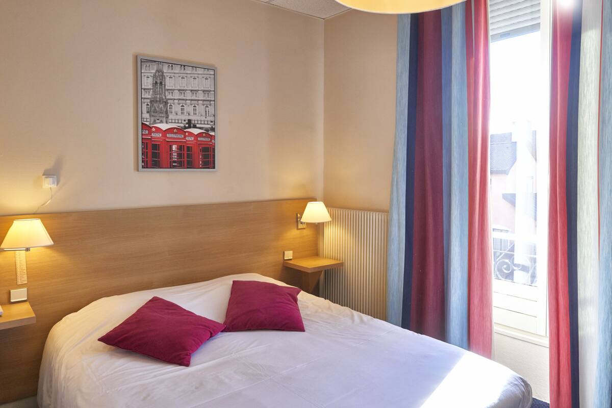 A hotel room with a bed, two bedside lamps, a wall-mounted picture, and a window with curtains.
