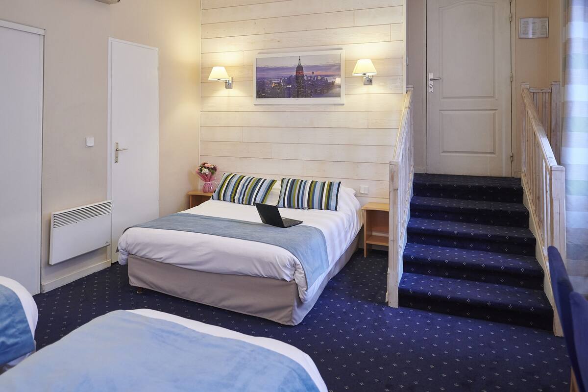 A hotel room with two beds, a laptop on one bed, a cityscape painting above the bed, and a staircase to the right.