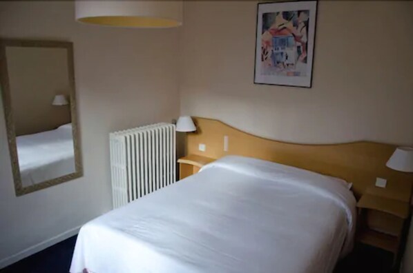 A hotel room with a bed, a nightstand, a lamp, a radiator, and a framed picture on the wall.