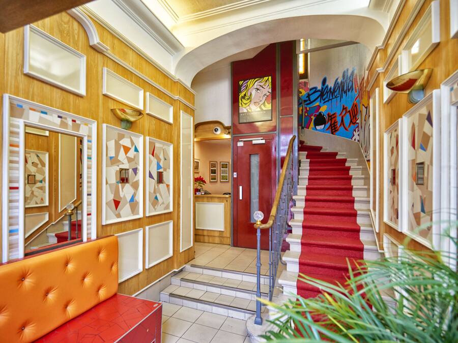 Un hall d’hôtel avec un escalier recouvert d’une moquette rouge, un comptoir d’accueil et un espace détente avec un banc rouge.