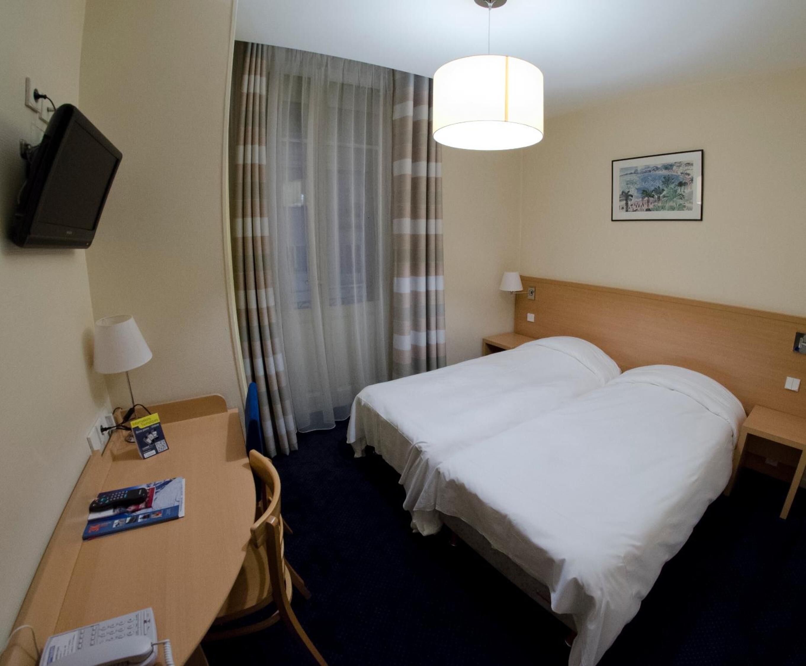 Habitación de hotel con cama, escritorio, silla, televisión, lámpara y ventana con cortinas.