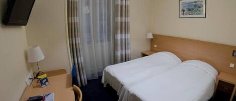 Habitación de hotel con cama, escritorio, silla, televisión, lámpara y ventana con cortinas.