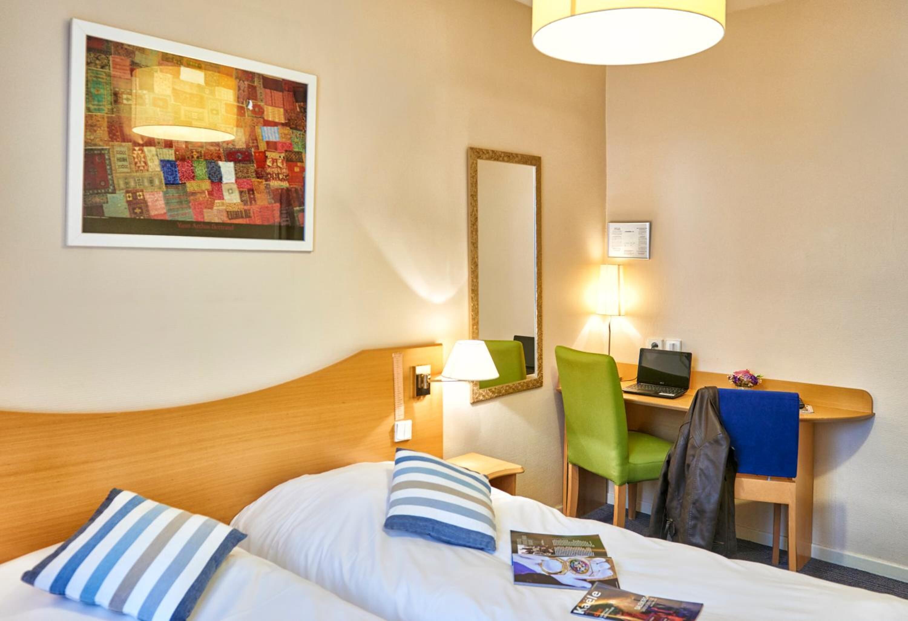 A hotel room with a bed, a desk with a laptop, a chair, a lamp, a mirror, and a framed artwork on the wall.