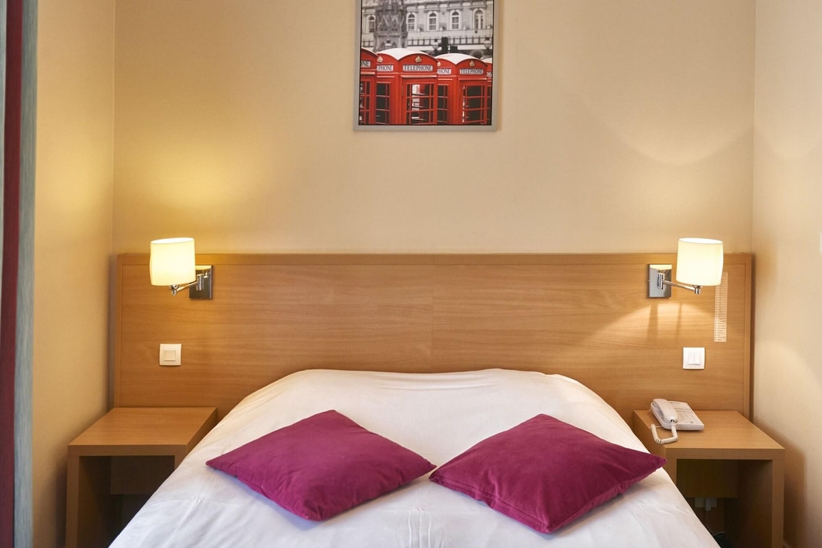 Una habitación de hotel con cabecera de madera, dos lámparas de noche, un teléfono y dos almohadas moradas.