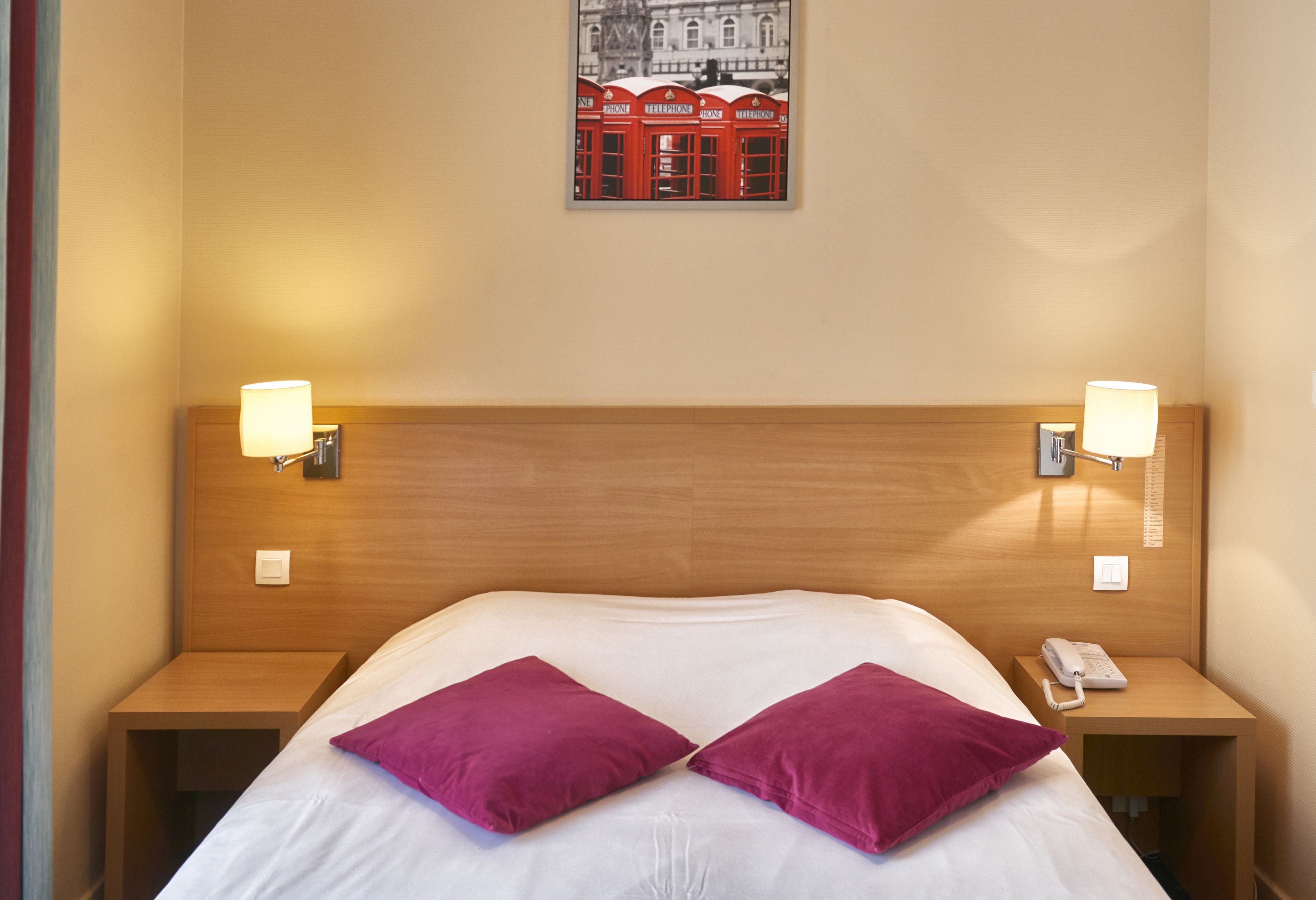 A hotel room with a wooden headboard, two bedside tables with lamps, a bed with white bedding and two purple pillows, and a framed picture on the wall.