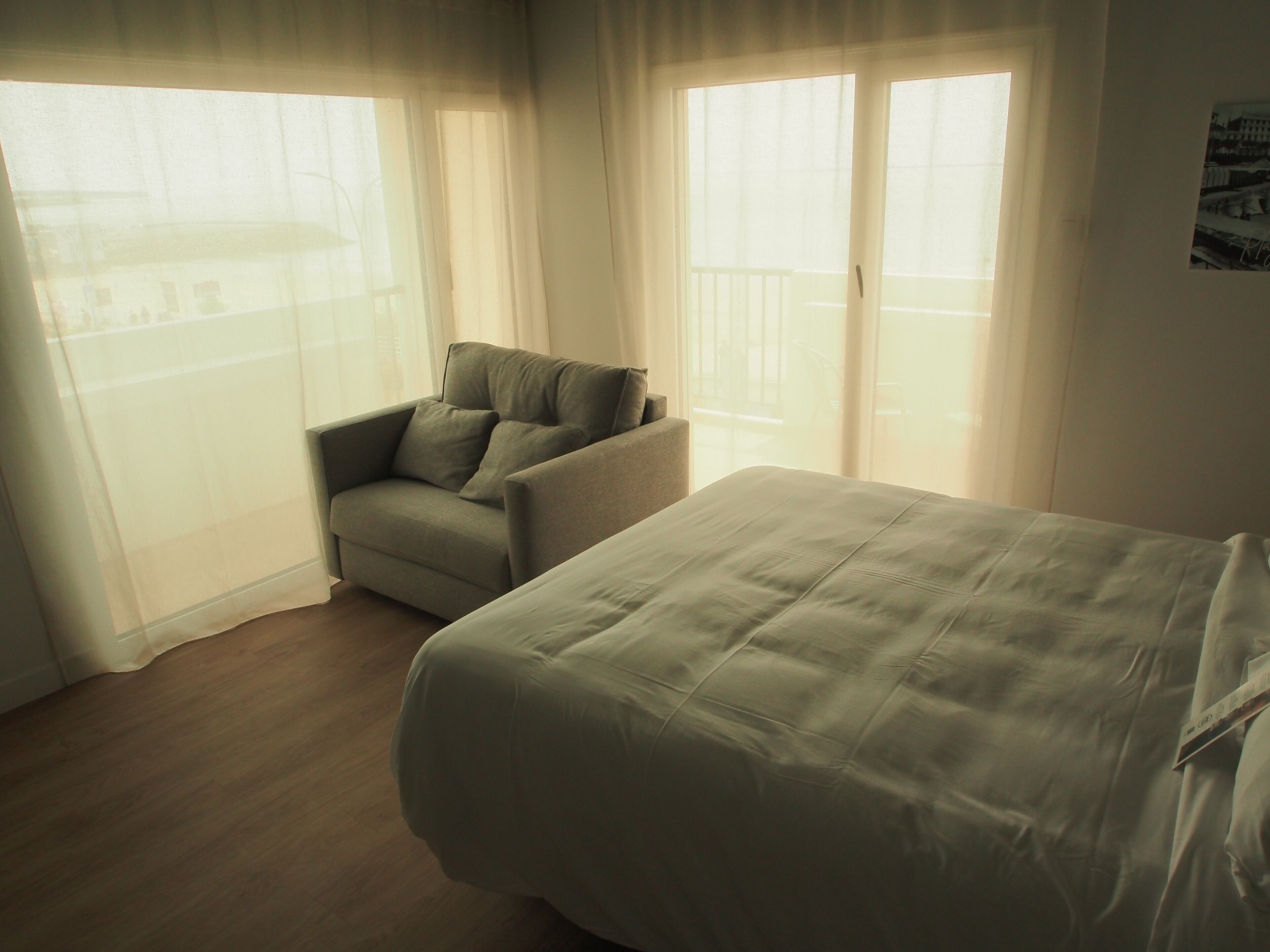 A room with a bed, a chair, and a balcony with curtains.
