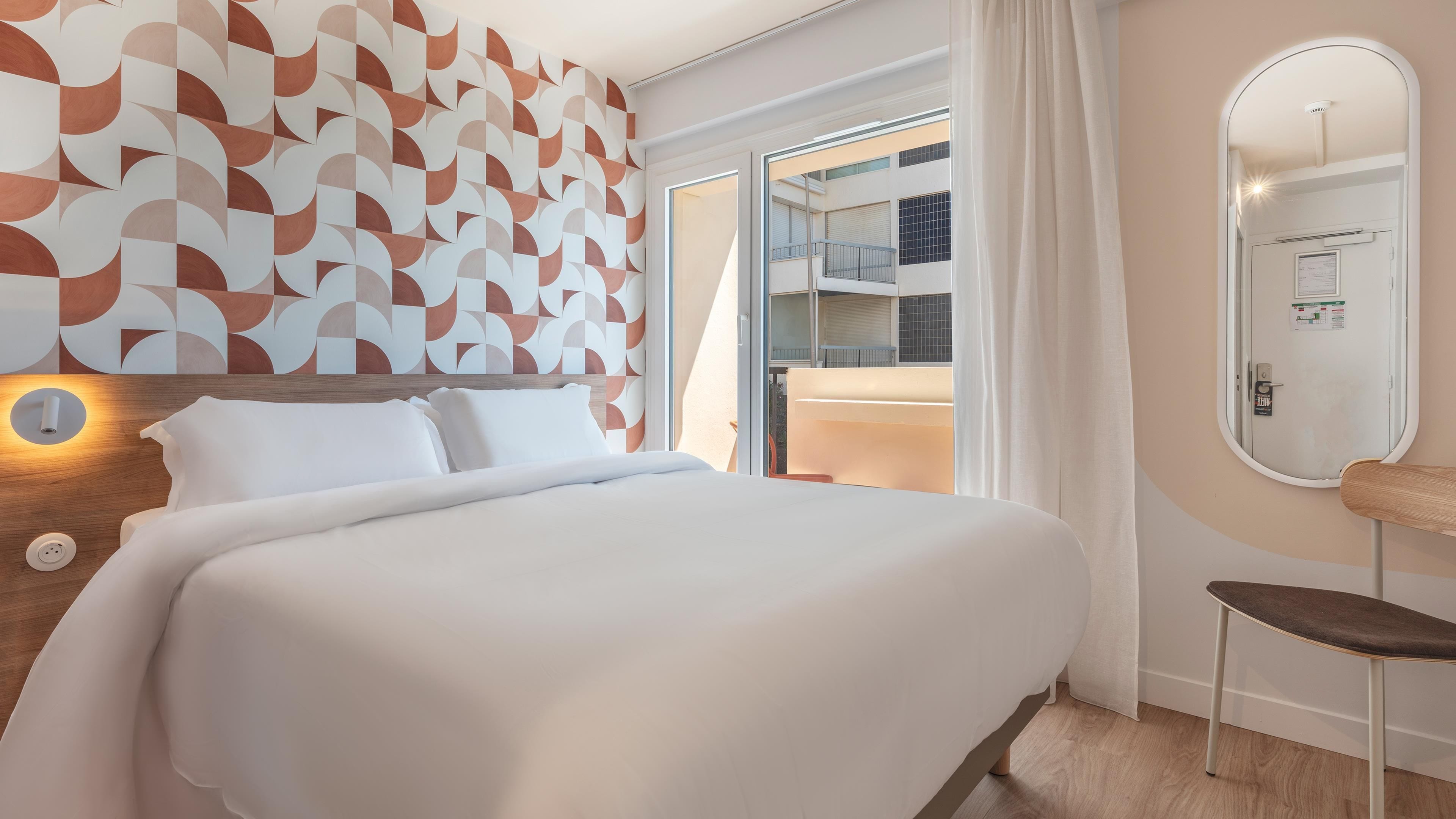 A hotel room with a bed, a wall with geometric patterns, a door leading to a balcony, and a mirror on the wall.