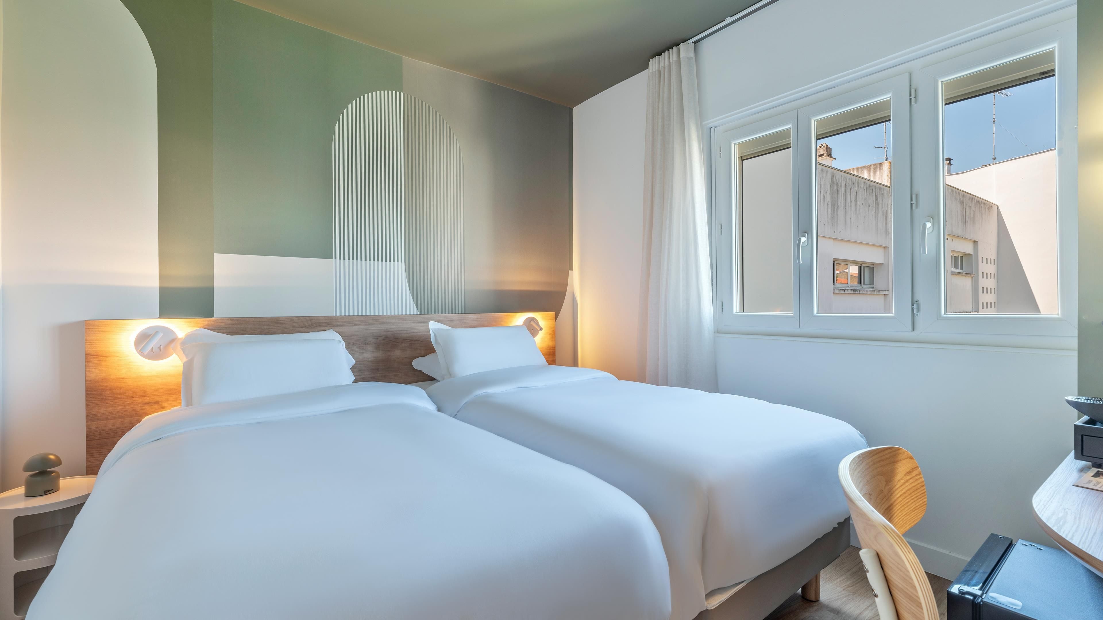 A hotel room with a double bed, bedside lamps, a desk with a chair, and a window with a view of buildings.