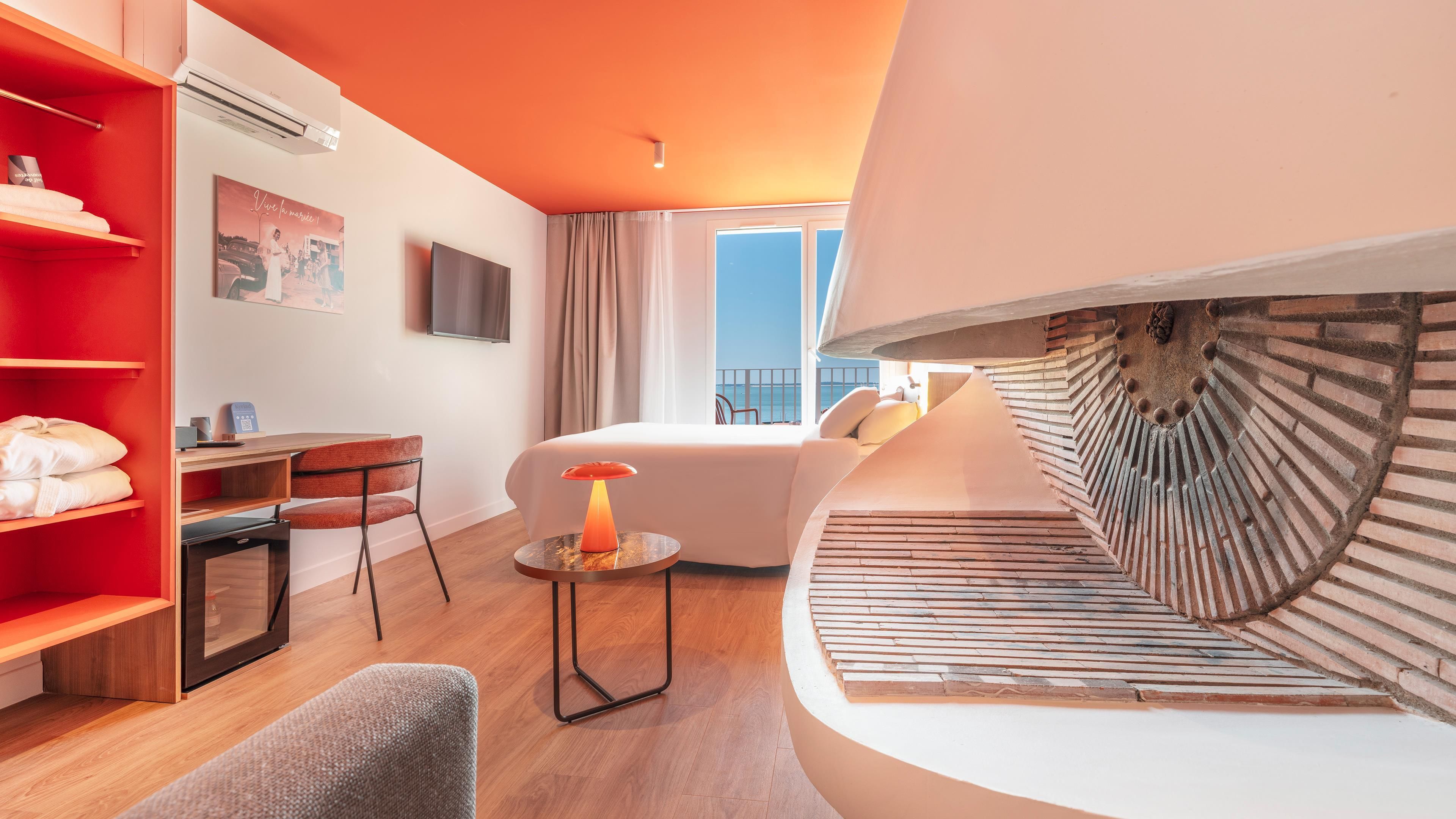 Une chambre d’hôtel moderne, équipée d’un lit, d’un bureau, d’une chaise, d’une petite table avec une lampe, d’une étagère rouge et d’une grande fenêtre donnant sur un beau paysage.
