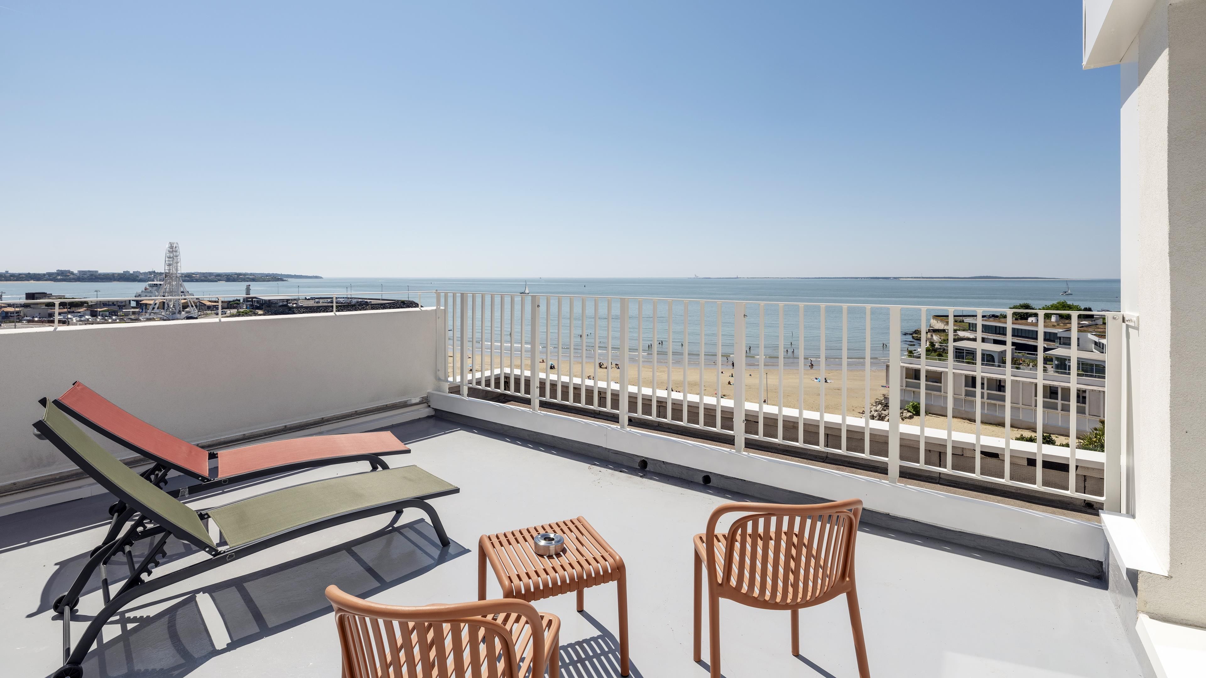 A balcony with a view of the beach, a lounge chair, a small table, and a chair.