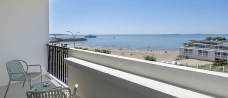 A balcony with two chairs and a table, overlooking a beach with people and a ship in the distance.