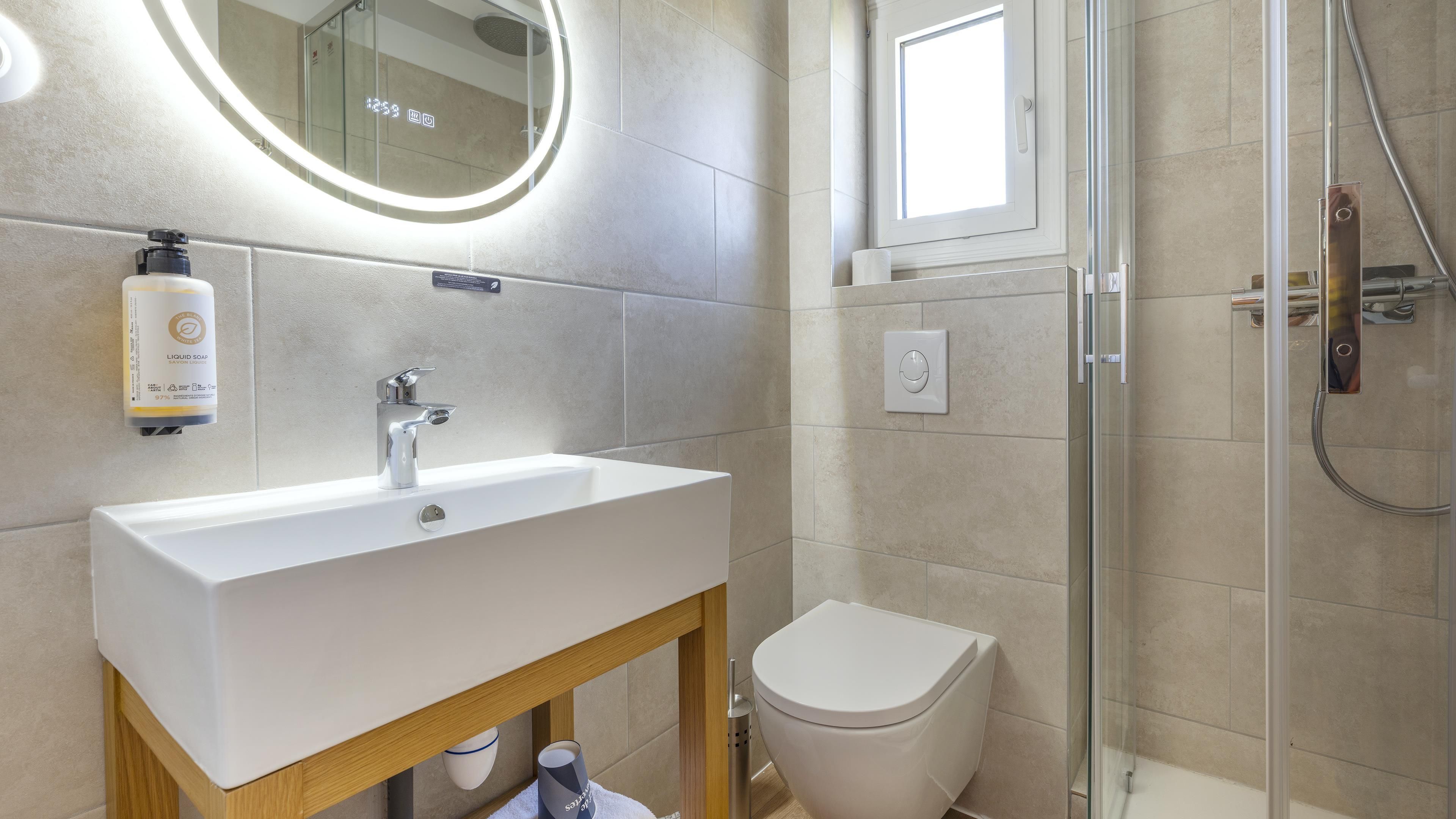 Une salle de bain moderne avec un lavabo blanc, un distributeur de savon manuel, des toilettes et une cabine de douche en verre.