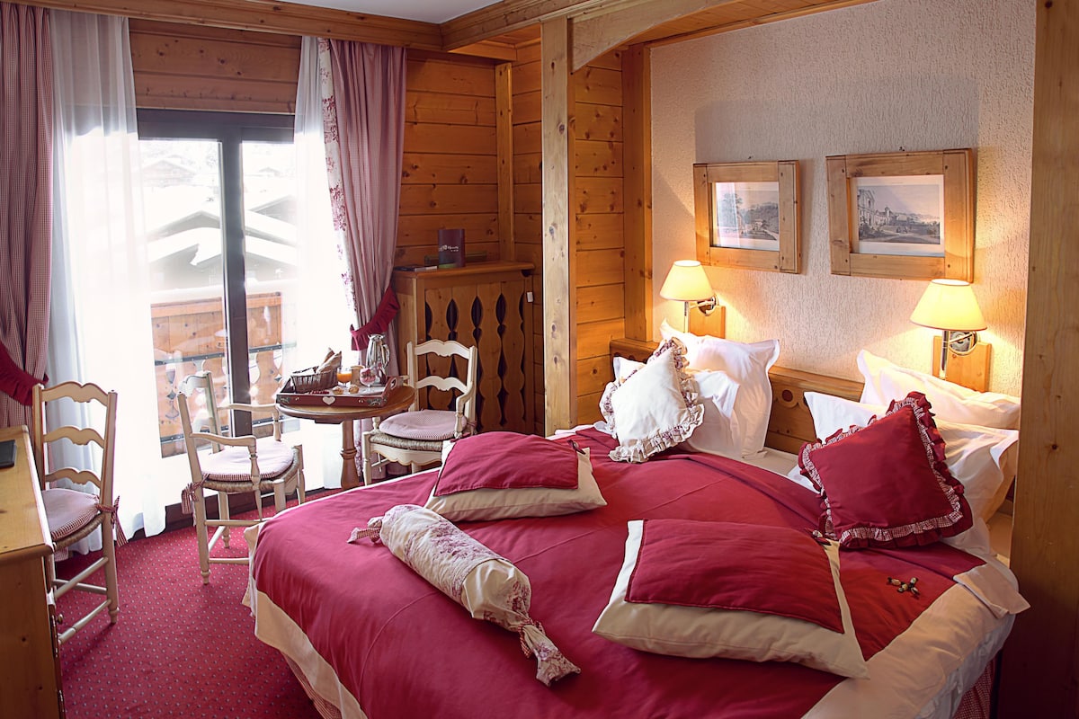 Una habitación tipo cabaña de madera con una cama grande, dos mesitas de noche con lámparas, una silla de madera y una pequeña mesa con una cesta.