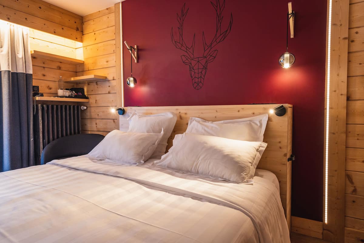 Una habitación de madera con una cama, un pared rojo decorado con un mural de cabeza de ciervo y dos lámparas fijadas a la pared.
