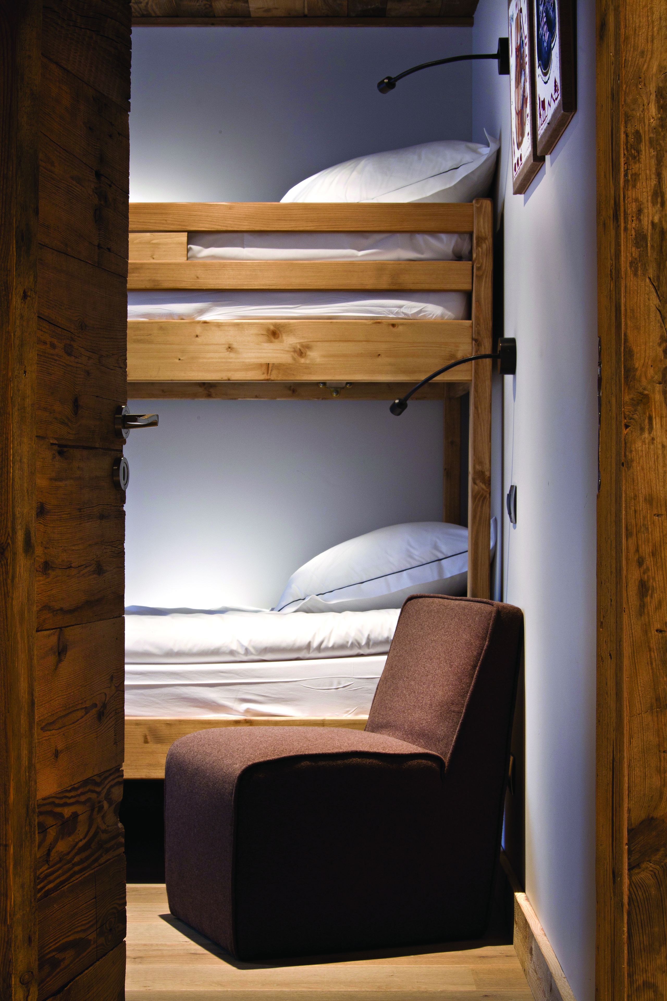 Una cama litera con cabecera de madera, una cama individual en la parte inferior y una silla marrón en la esquina.