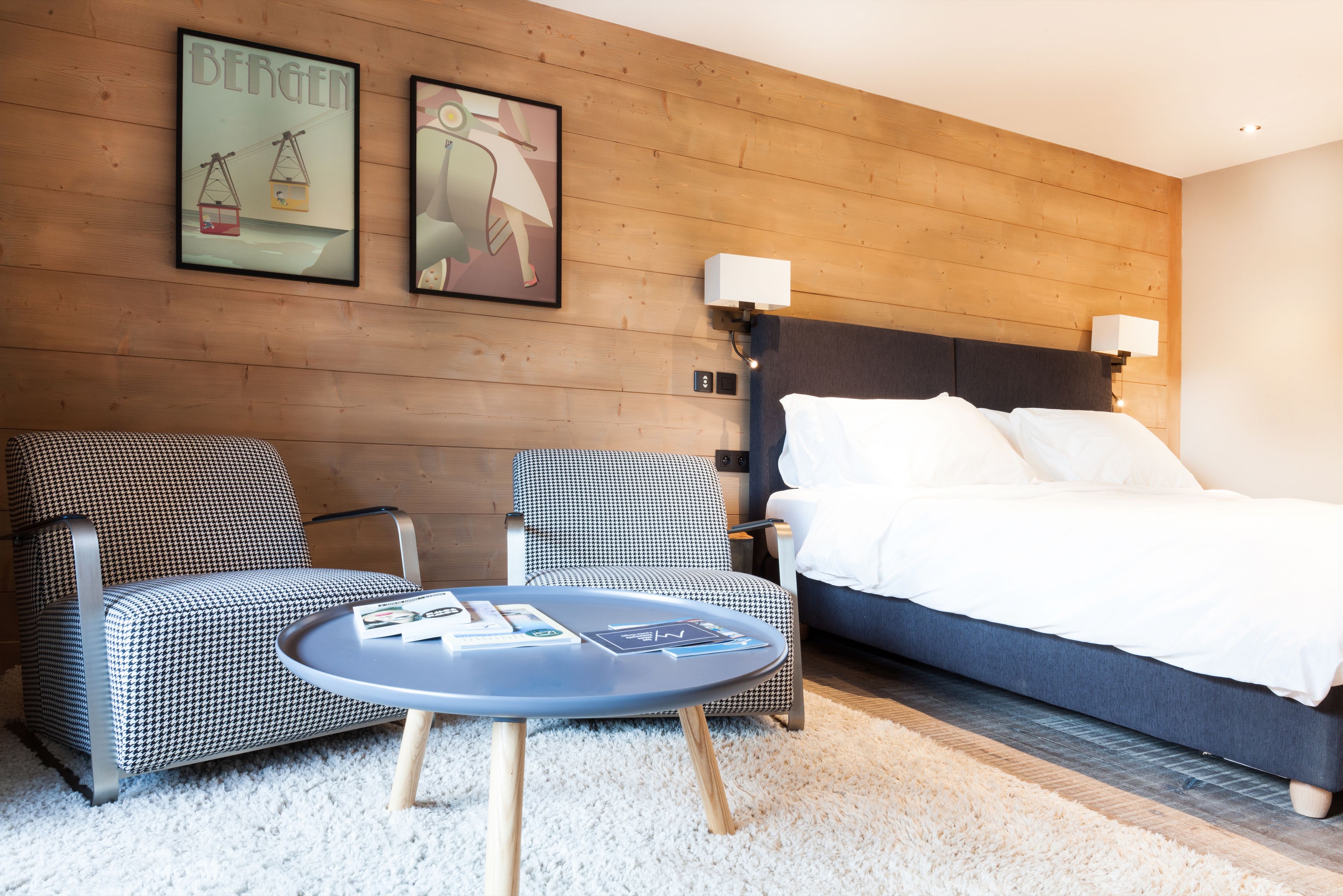 Quarto de hotel com parede de madeira, duas poltronas estampadas xadrez, uma mesa redonda com revistas, uma cama grande com lençóis brancos e duas luminárias fixadas na parede.
