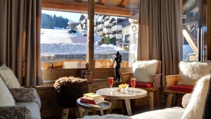 A cozy room with a wooden interior, large windows offering a view of a snowy mountain area, a statue, and a table with books and glasses.