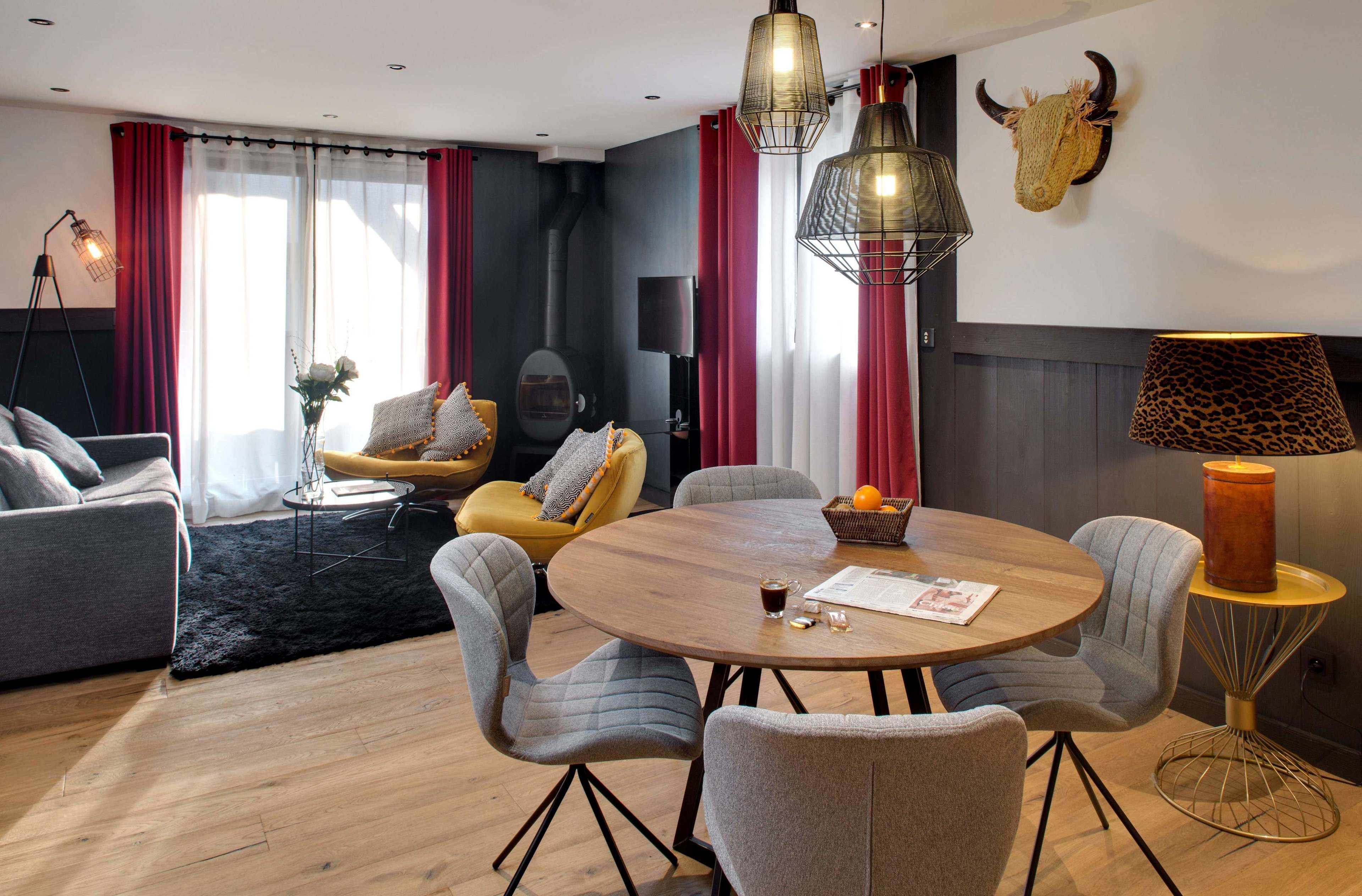 A modern living room with a round wooden table, grey upholstered chairs, a yellow armchair, and a red curtain.