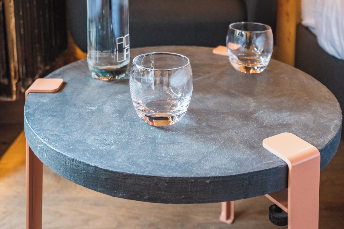 Una pequeña mesa redonda con una botella y dos vasos, una silla negra, y una pared de madera con un detalle rojo.