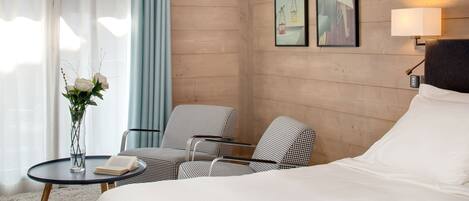 A hotel room with a bed, two chairs, a small table with a book and a vase of flowers, and two framed pictures on the wall.