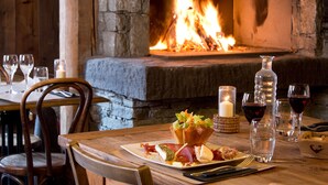 A dining table with a plate of food, glasses, and a candle. A fireplace with a fire is visible in the background.