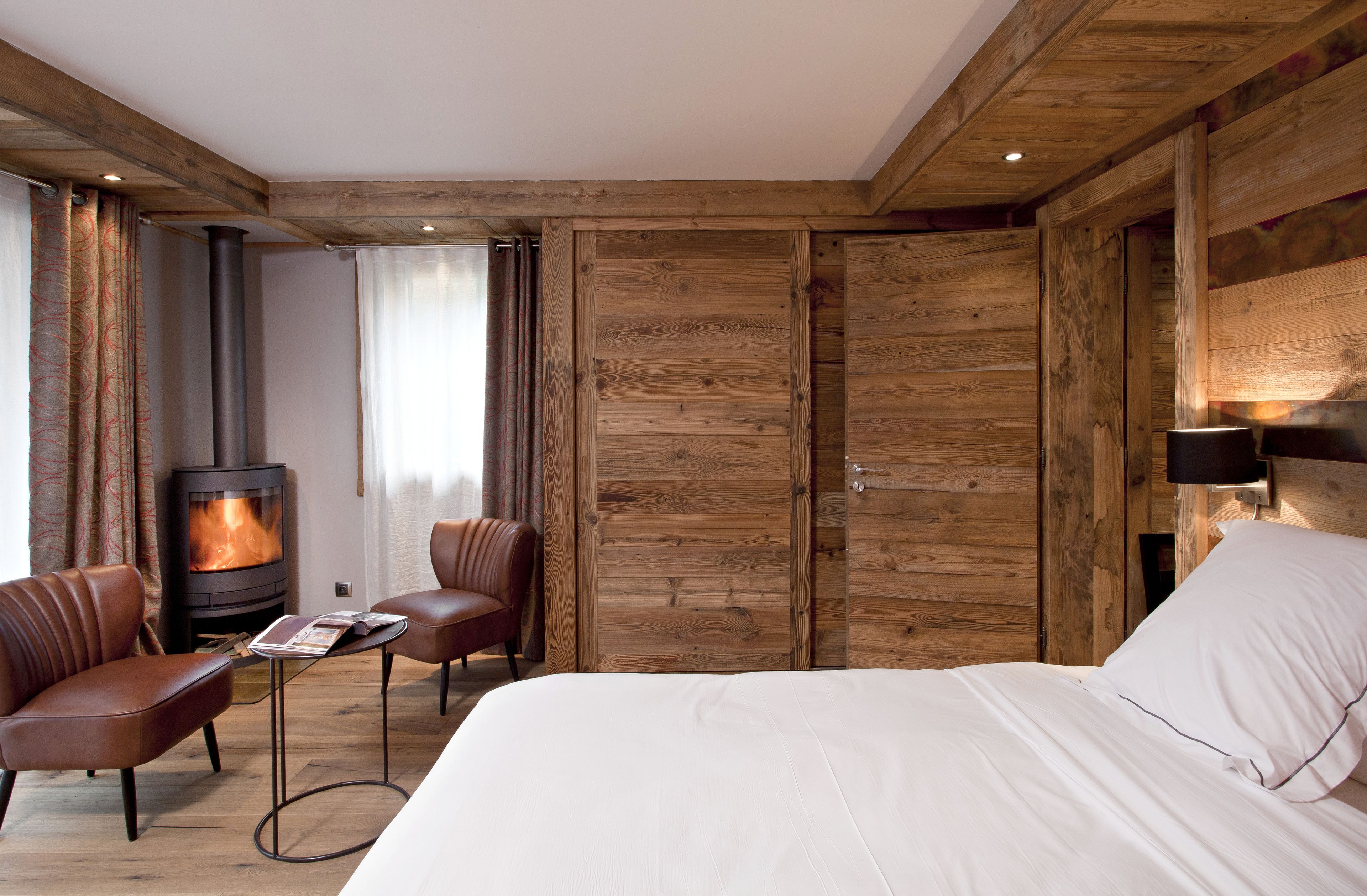 A cozy room with a bed, two leather chairs, a small table, a wood-paneled wall, and a wood-burning stove.