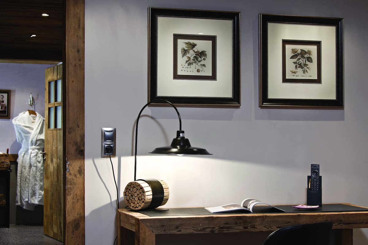 Una habitación con un escritorio de madera, una lámpara, una impresión enmarcadas de motivos botánicos y un control remoto.