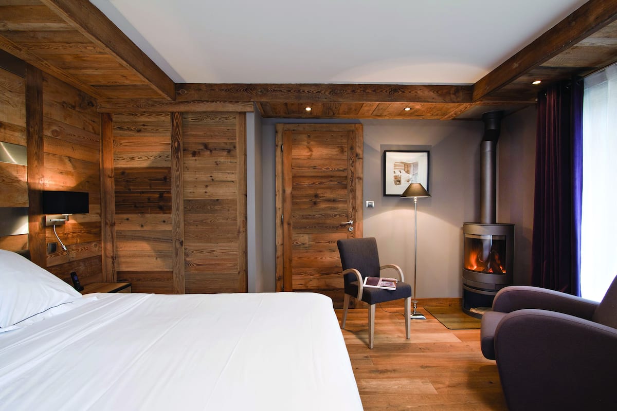 Habitación de hotel con interior de madera, una cama, un sillón, un sofá, una lámpara de pared y un estufa de leña.