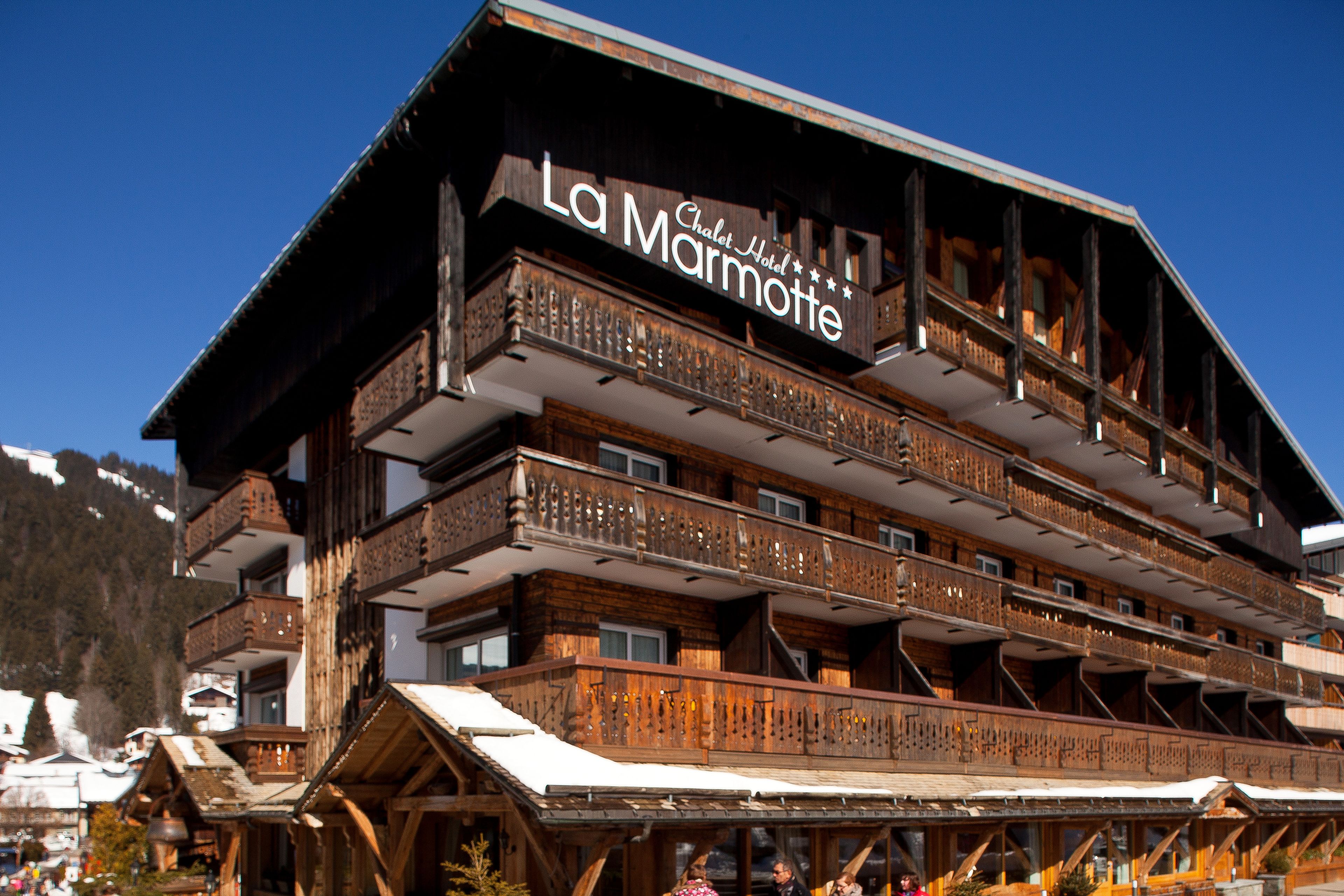 A multi-story wooden building with balconies and a sign that reads "La Marmotte Chalet Hotel."