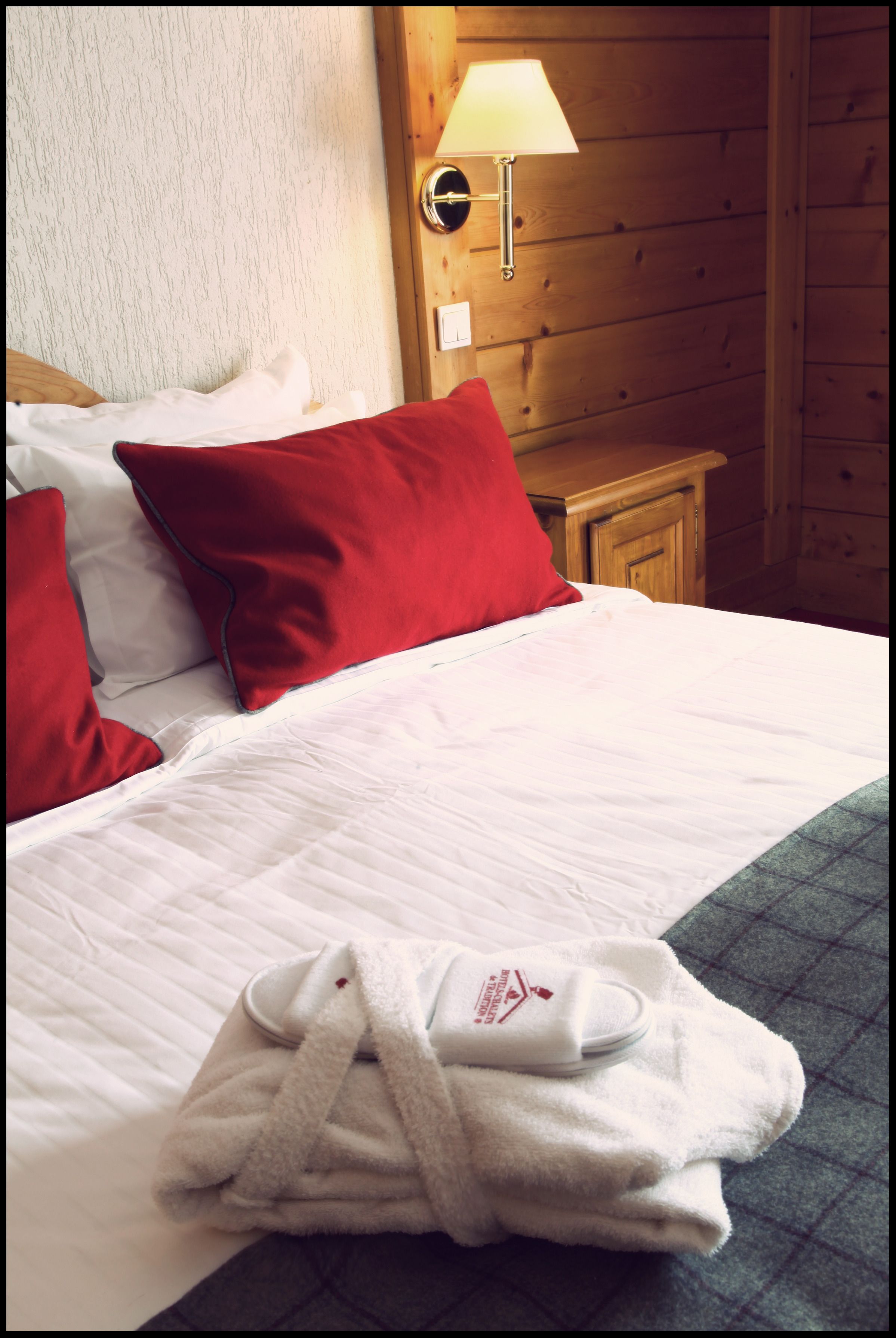 A neatly made bed with white and red pillows, a white bathrobe on the bed, and a wooden nightstand with a lamp.