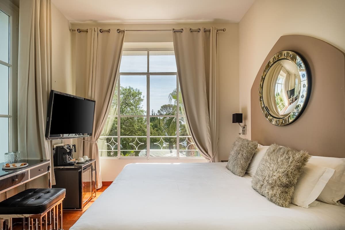 A hotel room with a large bed, a TV, a coffee maker, and a view of greenery outside.
