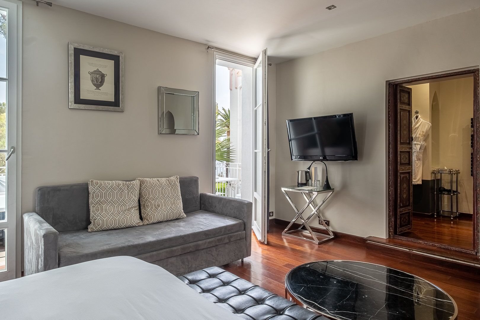 A modern hotel room with a grey sofa, a bed, a TV, and a small table with a lamp.