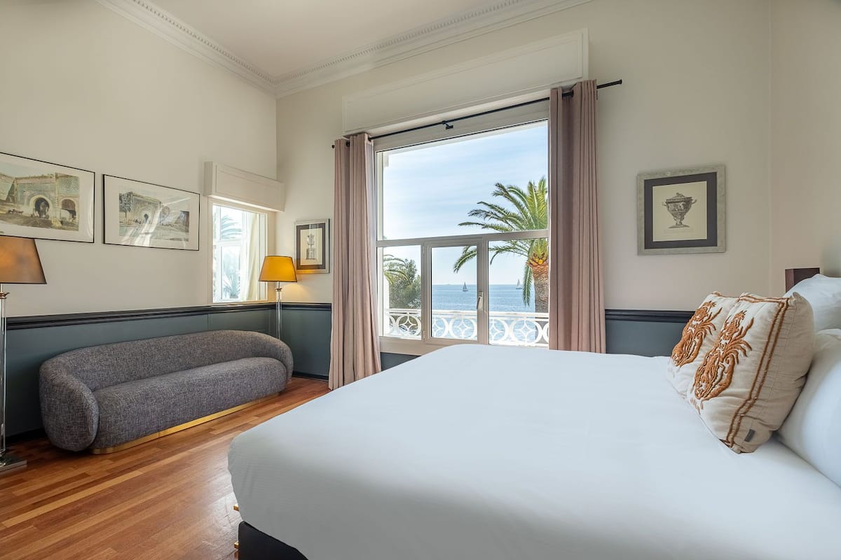 A hotel room with a large bed, a sofa, a balcony with a view of the sea, and framed pictures on the walls.