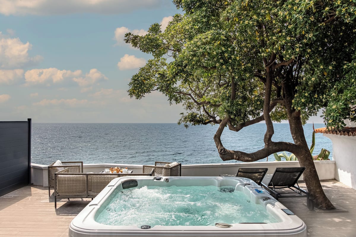 A hot tub with a view of the sea, surrounded by lounge chairs and a tree.