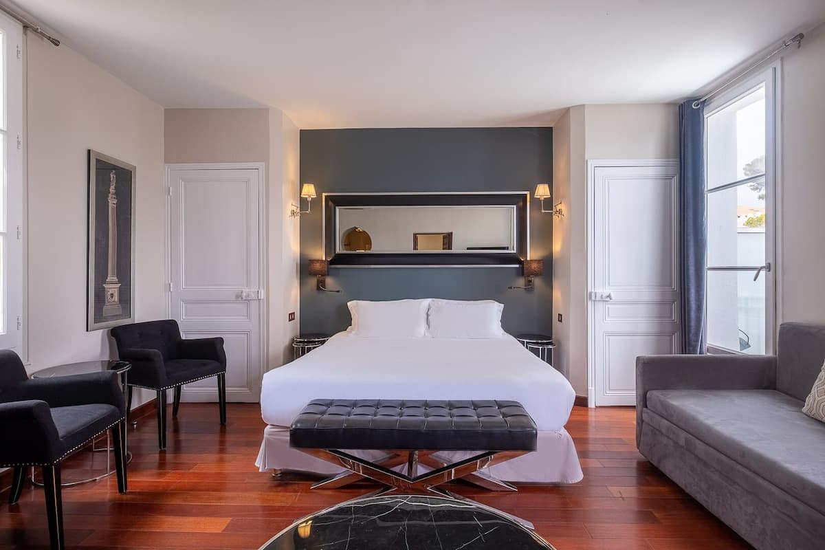 A modern hotel room with a large bed, a sofa, two armchairs, a round coffee table, and a large window with curtains.
