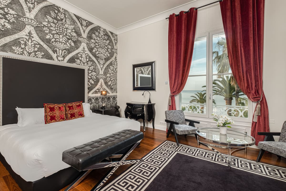 A bedroom with a large bed, a small table, two chairs, a window with red curtains, and a patterned wall.