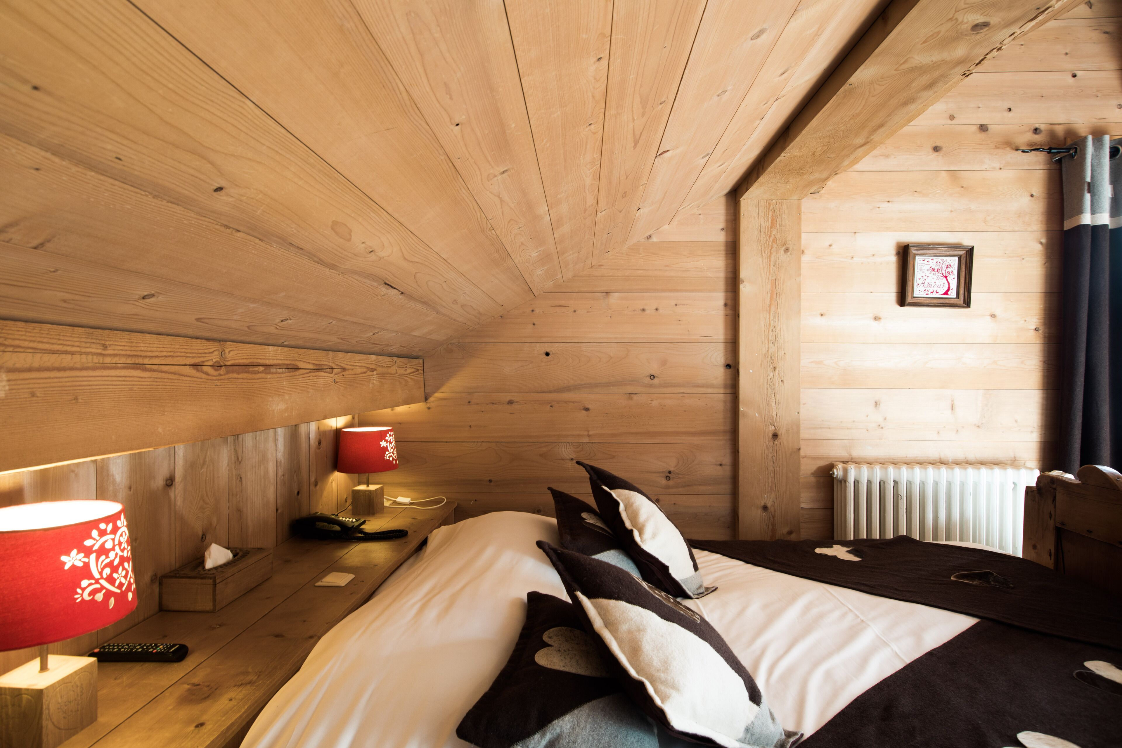 Habitación de cabaña de madera con una cama, una lámpara roja, un cuadro en la pared y una ventana con cortinas.