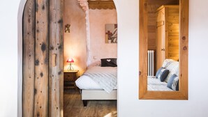 A bedroom with a wooden door, a bed with pillows, a nightstand with a lamp, and a wall-mounted mirror. - Hôtel La Griyotire (Praz-sur-Arly)