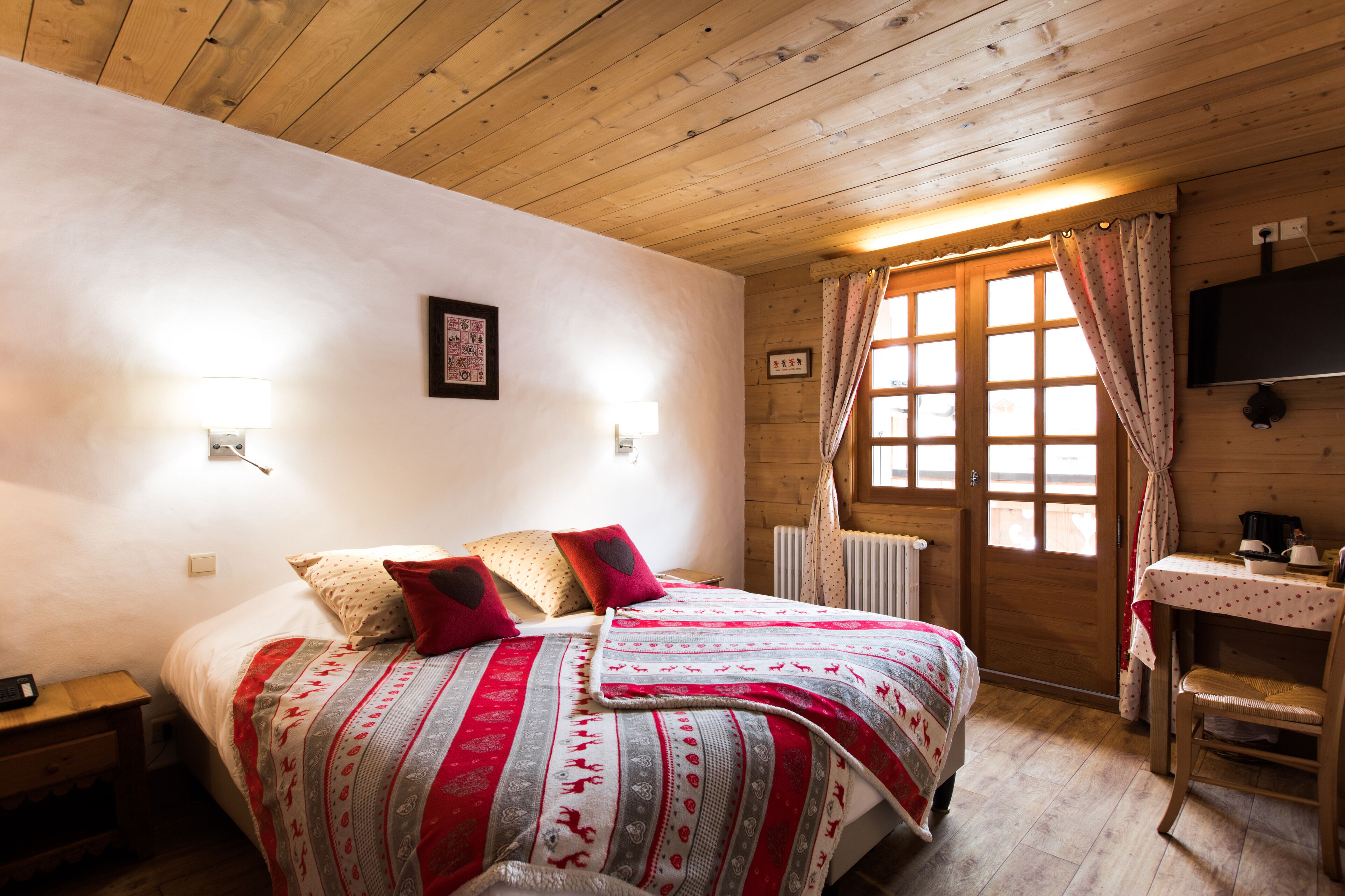 A cozy room with a wooden ceiling, a bed with a patterned blanket, a small table with a kettle, and a window with curtains.