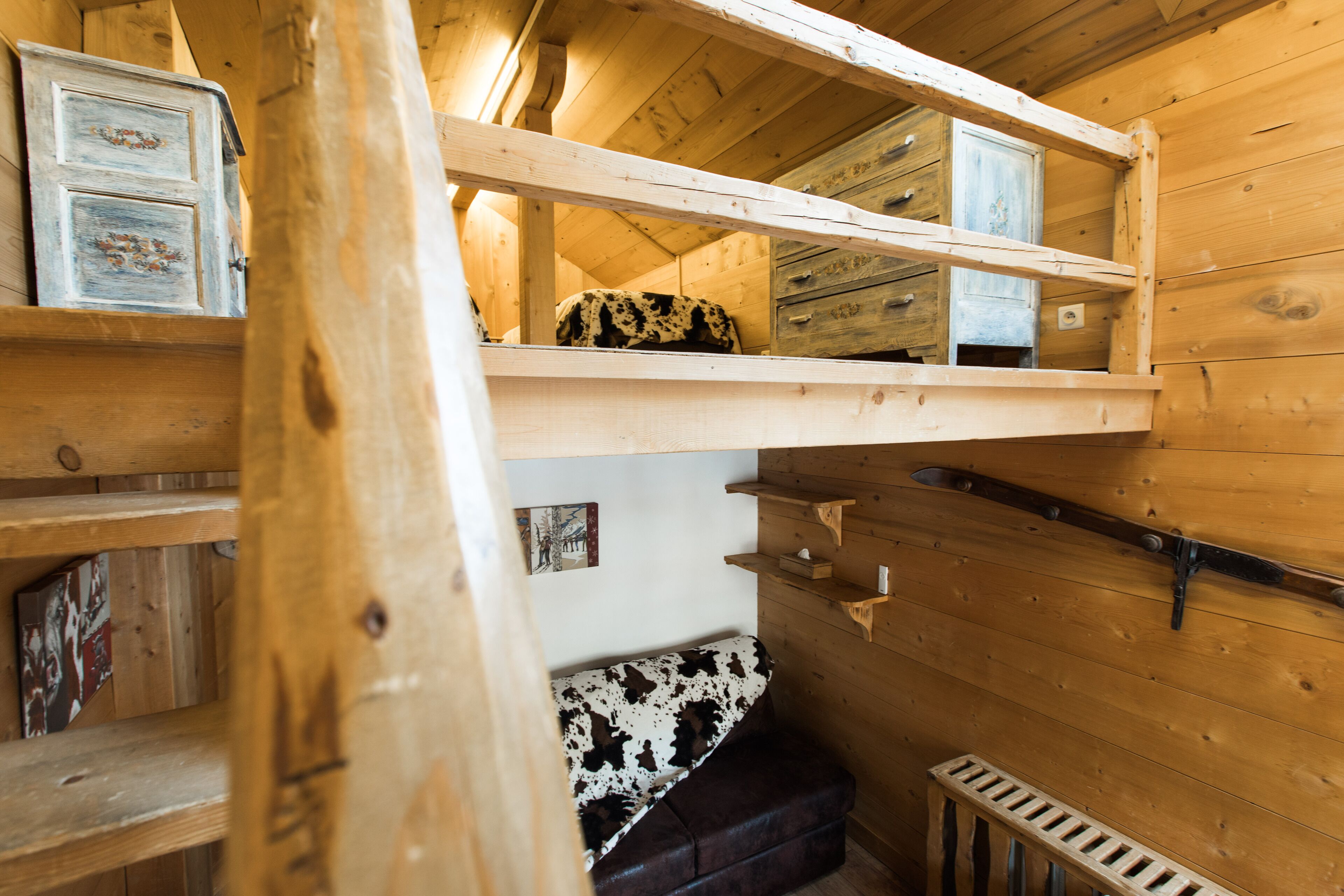 L’intérieur d’un chalet en bois est aménagé avec un lit superposé, un banc recouvert de peau de vache et des étagères en bois.