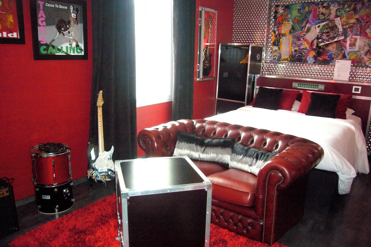 A bedroom with a red leather sofa, a drum set, and a bed with a white comforter.