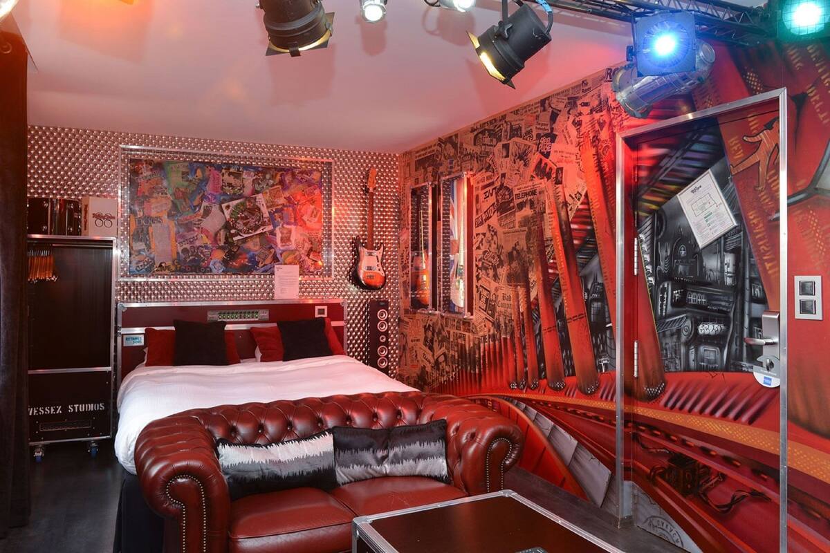 A room with a bed, a red leather sofa, a guitar, and a wall covered with posters and graffiti.