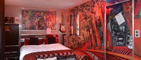 Una habitación con una cama, un sofá de cuero rojo, una guitarra y una pared cubierta de carteles y grafitis.