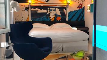 A modern hotel room with a bed, a blue chair, a glass coffee table, and a mural on the wall.