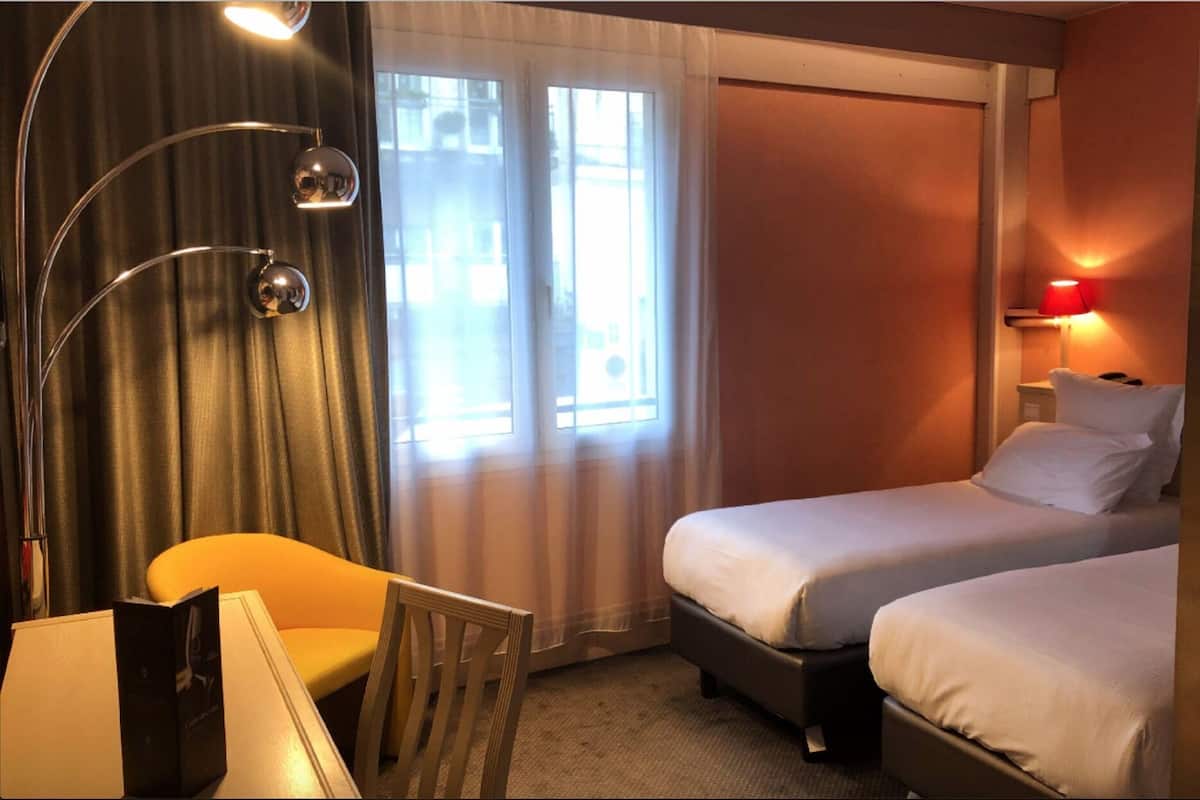 A hotel room with two beds, a desk, a chair, a lamp, and a window with curtains.